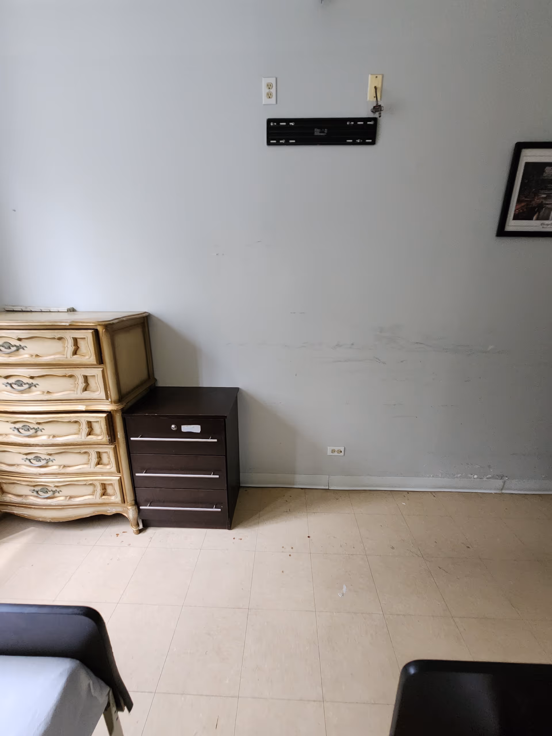 A sparsely furnished bedroom shows two dressers, a bed frame, and framed wall art.