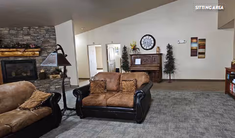 A sitting area in an assisted living facility featuring two cushioned armchairs with brown and black upholstery, a stone fireplace with a wooden mantel, a floor lamp, a piano against the far wall with decorative items on top, two tall artificial plants, a large wall clock, and framed artwork on the wall. The floor is carpeted and the space appears cozy and inviting.
