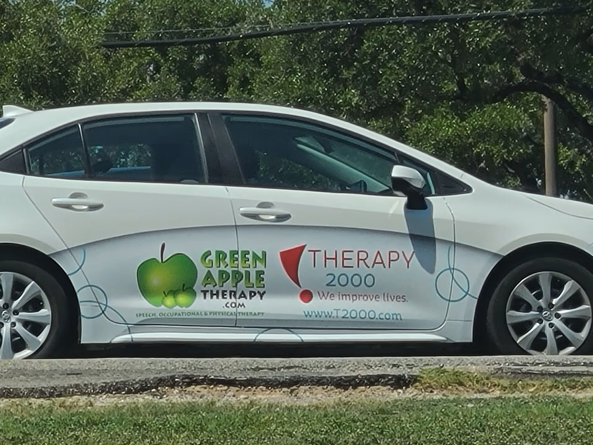 White sedan with Therapy 2000 and Green Apple Therapy logos parked outdoors.