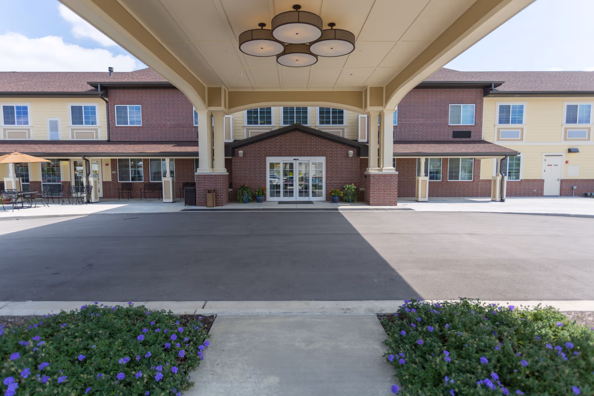 View of the front entrance of Meadow Brook Senior Living facility showing a covered driveway with a large overhead light fixture, brick and yellow siding exterior walls, glass double doors, outdoor seating area with tables and umbrellas, and flower beds with purple flowers in the foreground.