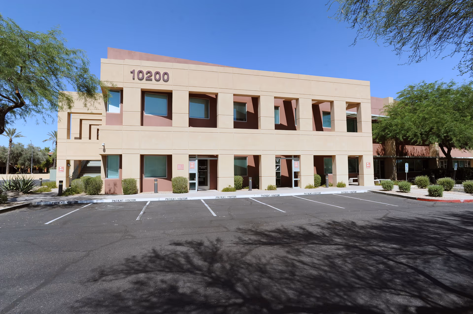 Two-story beige building with a large parking lot and desert landscaping in front.