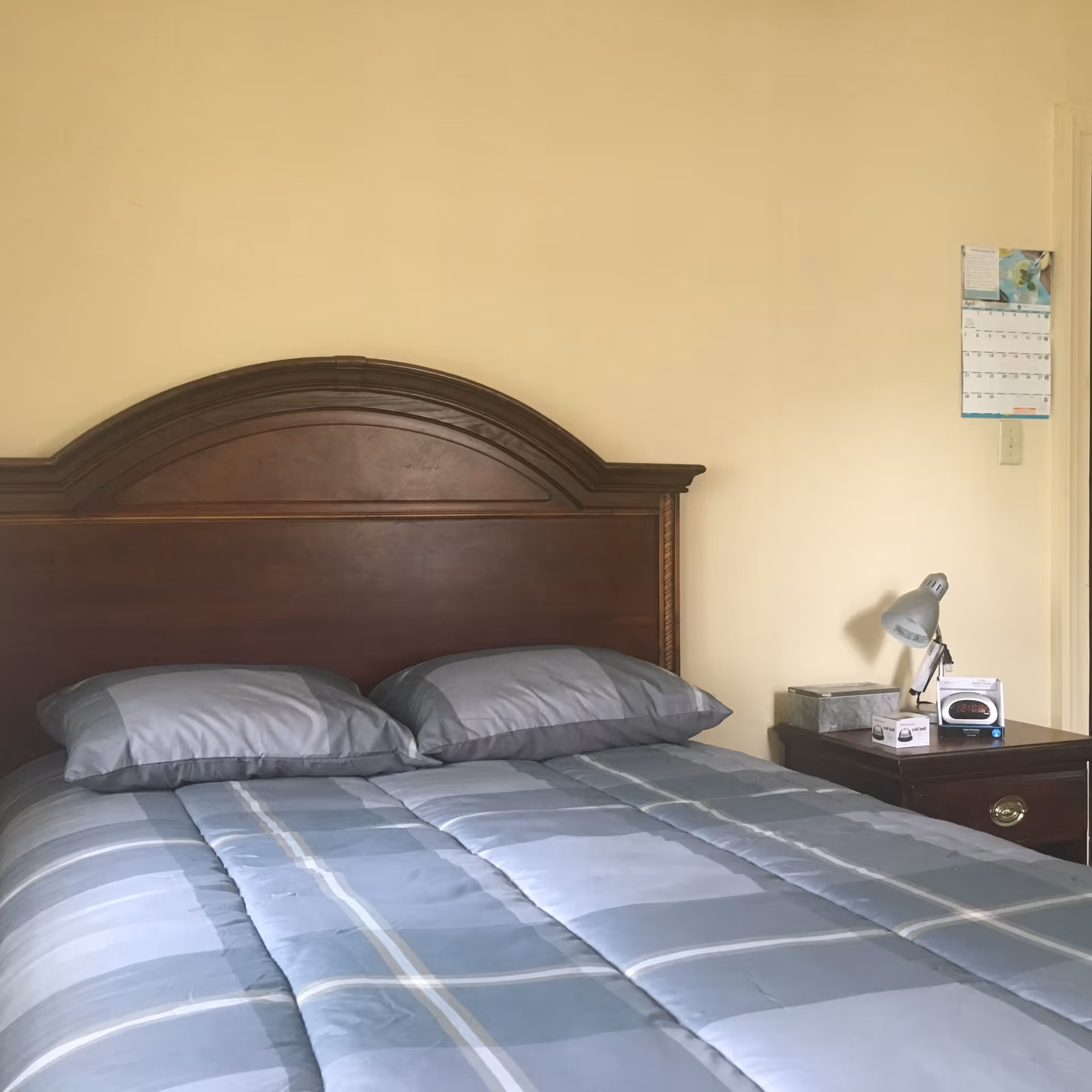 A bedroom with a wooden headboard bed made with a blue and gray striped comforter and two matching pillows. Next to the bed is a wooden nightstand with a silver desk lamp, a tissue box, a small clock, and a calendar hanging on the beige wall above.