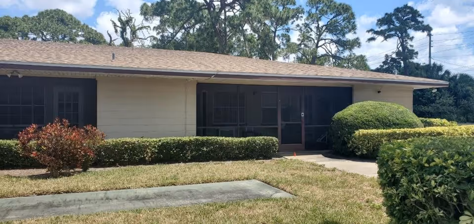 Single-story building with beige walls and a brown shingled roof, surrounded by neatly trimmed bushes and a small lawn under a partly cloudy sky.