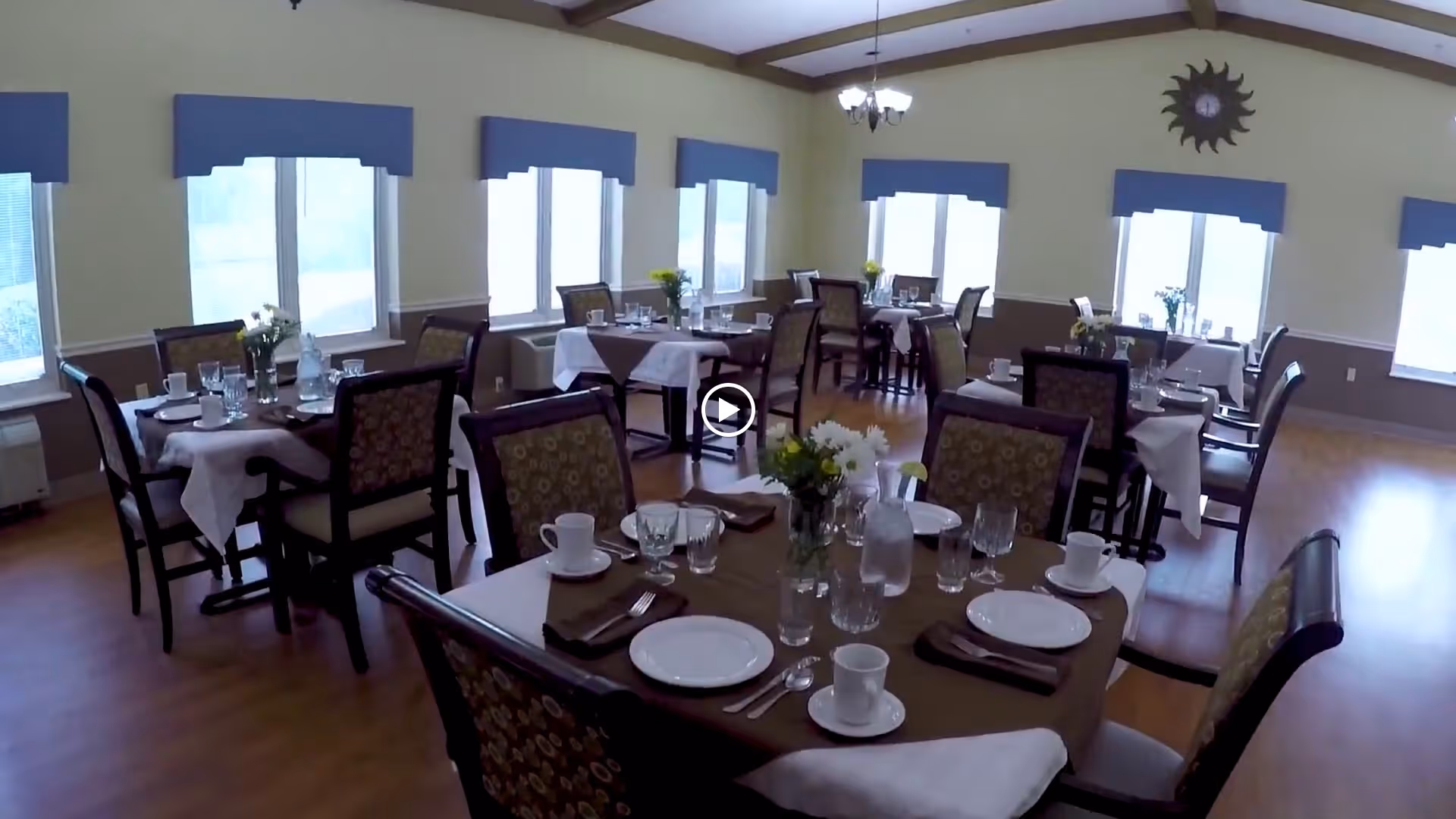 A dining room with multiple tables set for a meal. Each table has white plates, cups, glasses, silverware, and napkins. There are floral centerpieces on each table. The room has large windows with blue valances, wooden floors, and a ceiling with exposed beams and a chandelier. A sun-shaped clock is mounted on the wall.