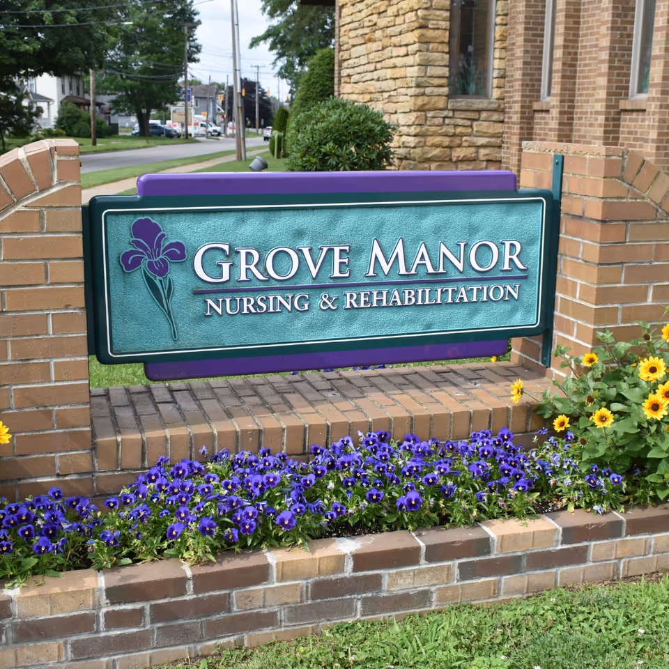 A brick entrance sign that reads "Grove Manor Nursing & Rehabilitation" above a flowerbed of purple pansies in front of a brick building.