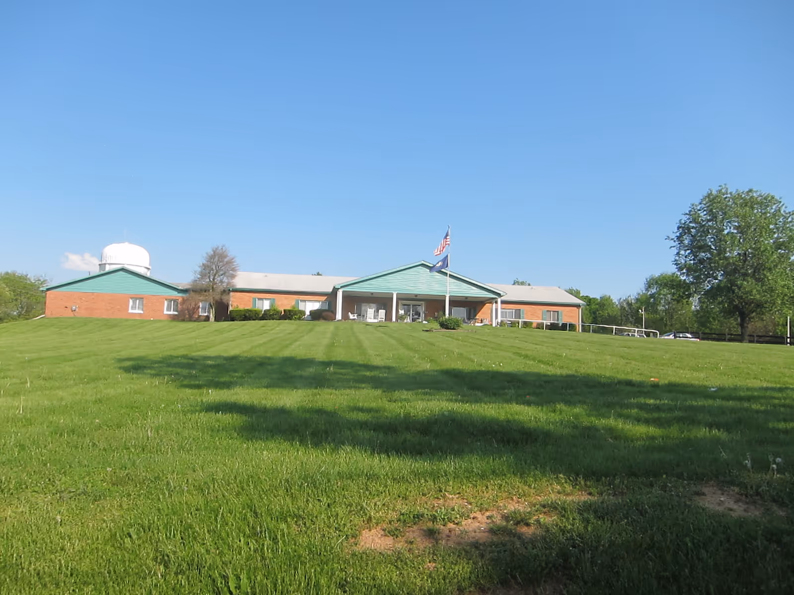 Wide view of a single-story brick building with a green roof and a covered entrance. Two flagpoles with American and state flags stand in front of the building. The building is surrounded by a large, well-maintained grassy lawn with trees in the background under a clear blue sky.