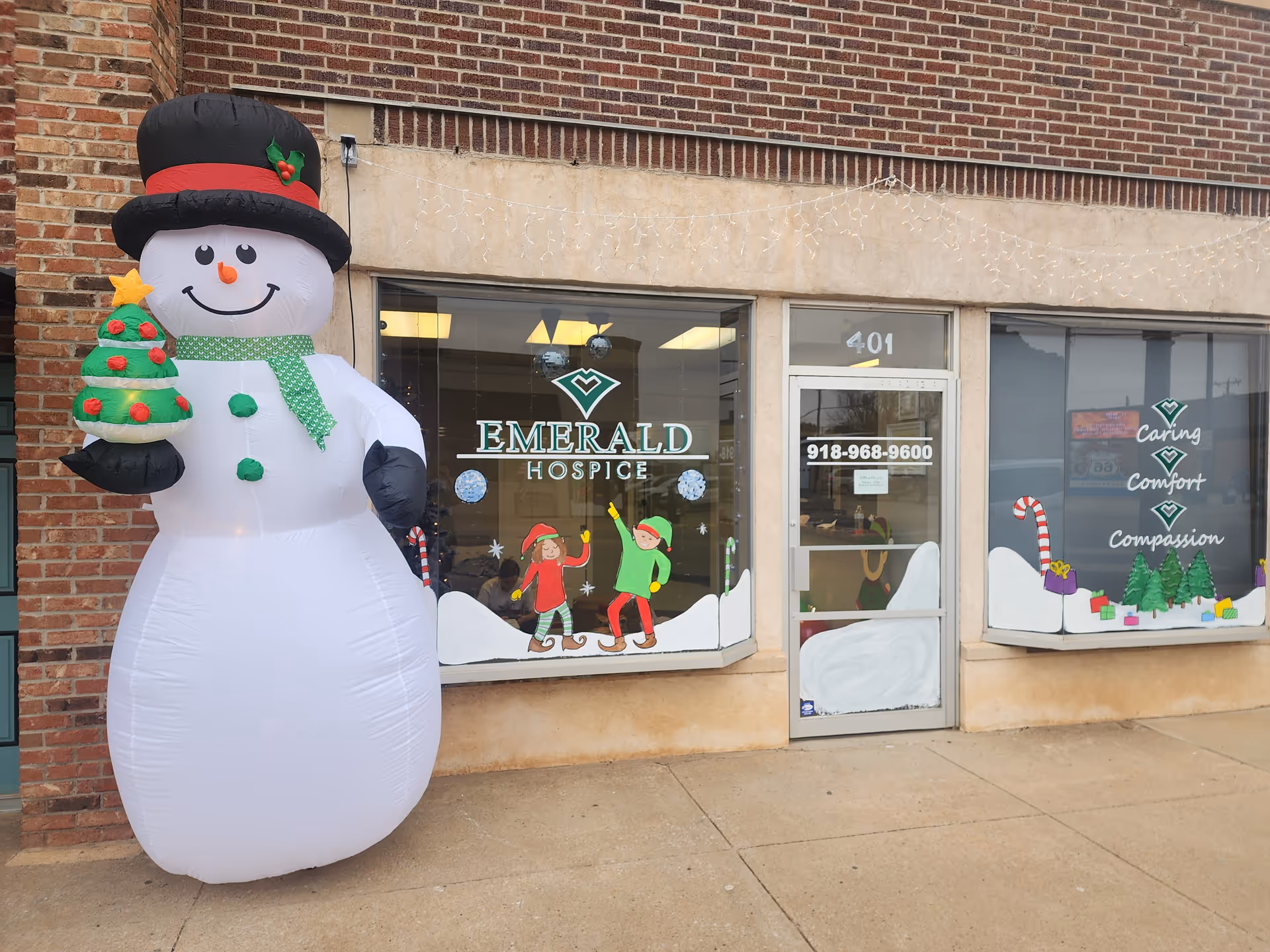 A large inflatable snowman stands outside Emerald Hospice storefront with holiday window decals.