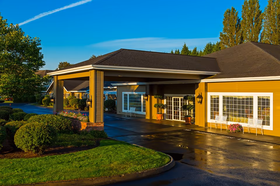 Exterior view of Canterbury Gardens Memory Care Community building entrance with a covered drop-off area, well-maintained landscaping including bushes and trees, and white benches near large windows under a clear blue sky.