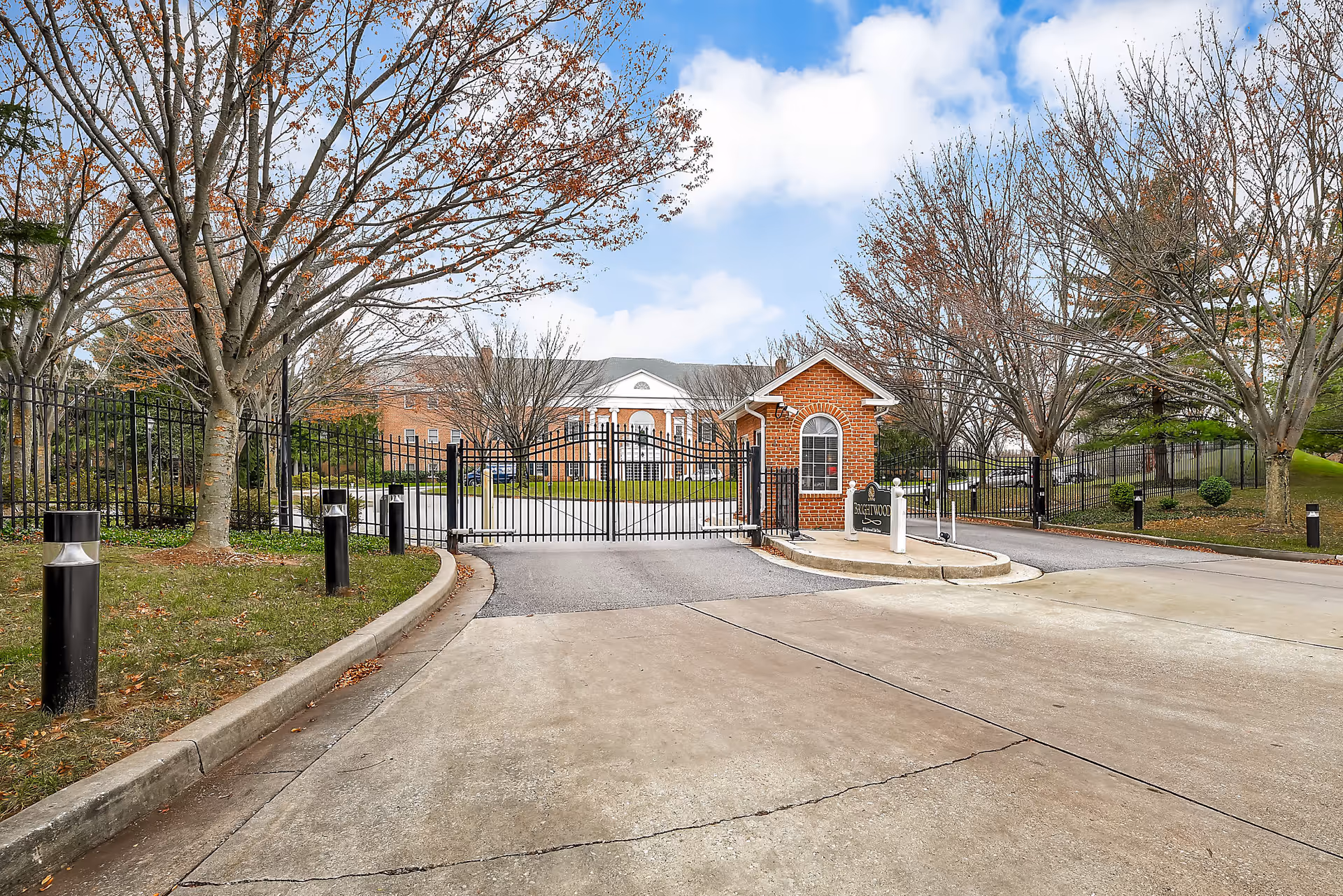Entrance gate to Brightwood Club Association with a brick guardhouse on the right side, black metal fencing, leafless trees, and a large brick building in the background under a partly cloudy sky.