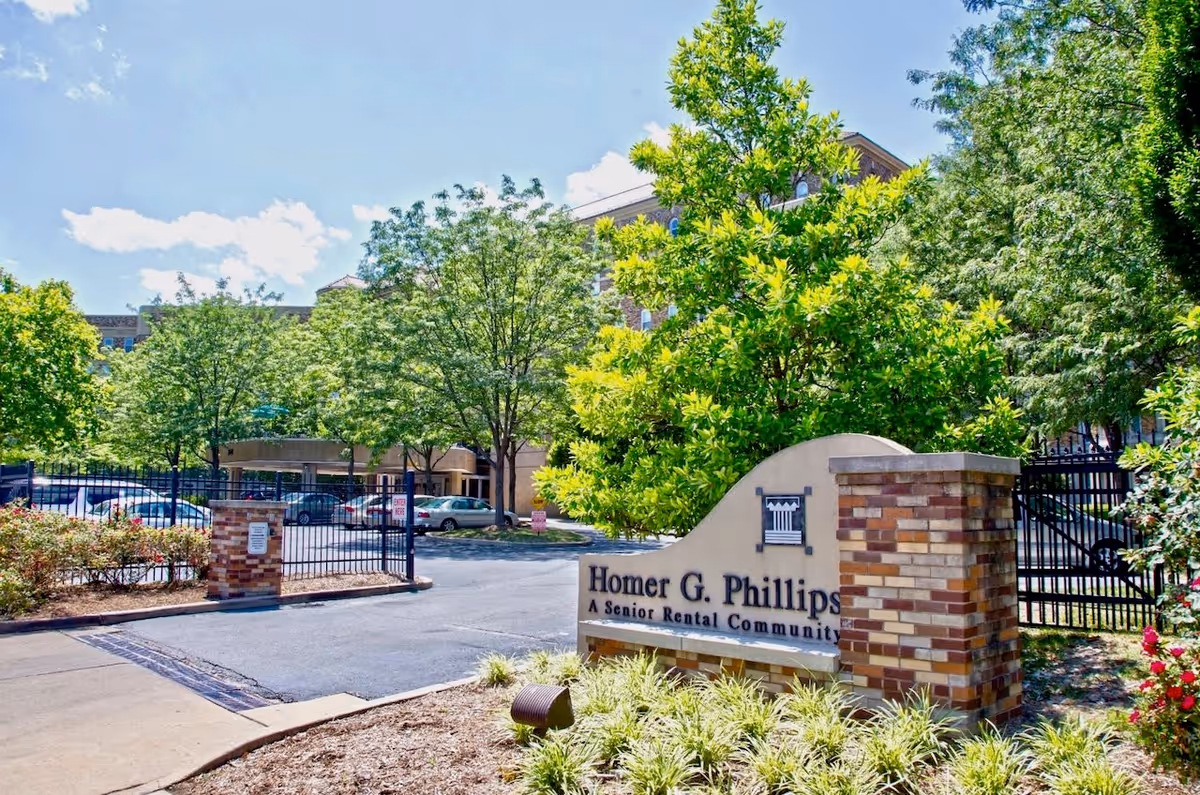Gated driveway and entrance sign for Homer G. Phillips senior apartments with trees and the building visible behind.