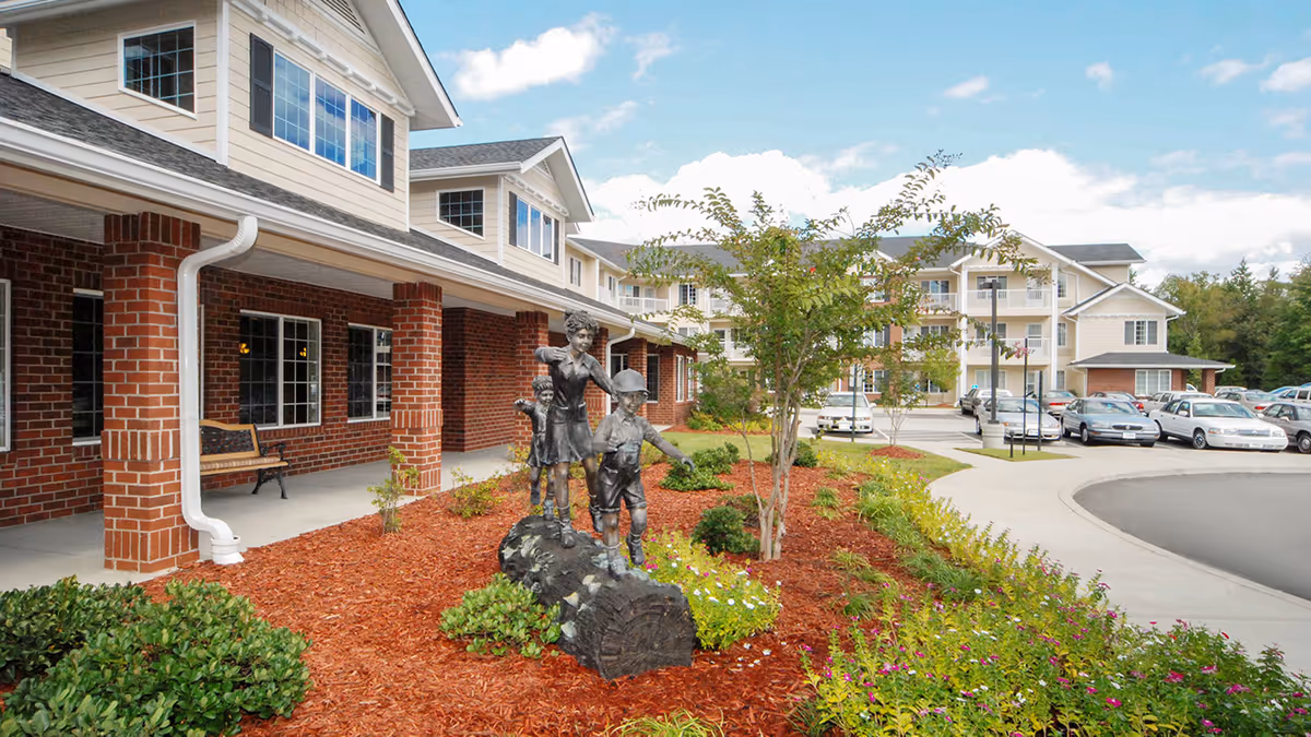 Exterior view of Dogwood Terrace by Barclay House featuring a brick and beige building with multiple windows and a covered walkway supported by brick columns. In the landscaped garden area, there is a bronze statue of three children playing on a log. Several cars are parked in the parking lot under a partly cloudy sky.
