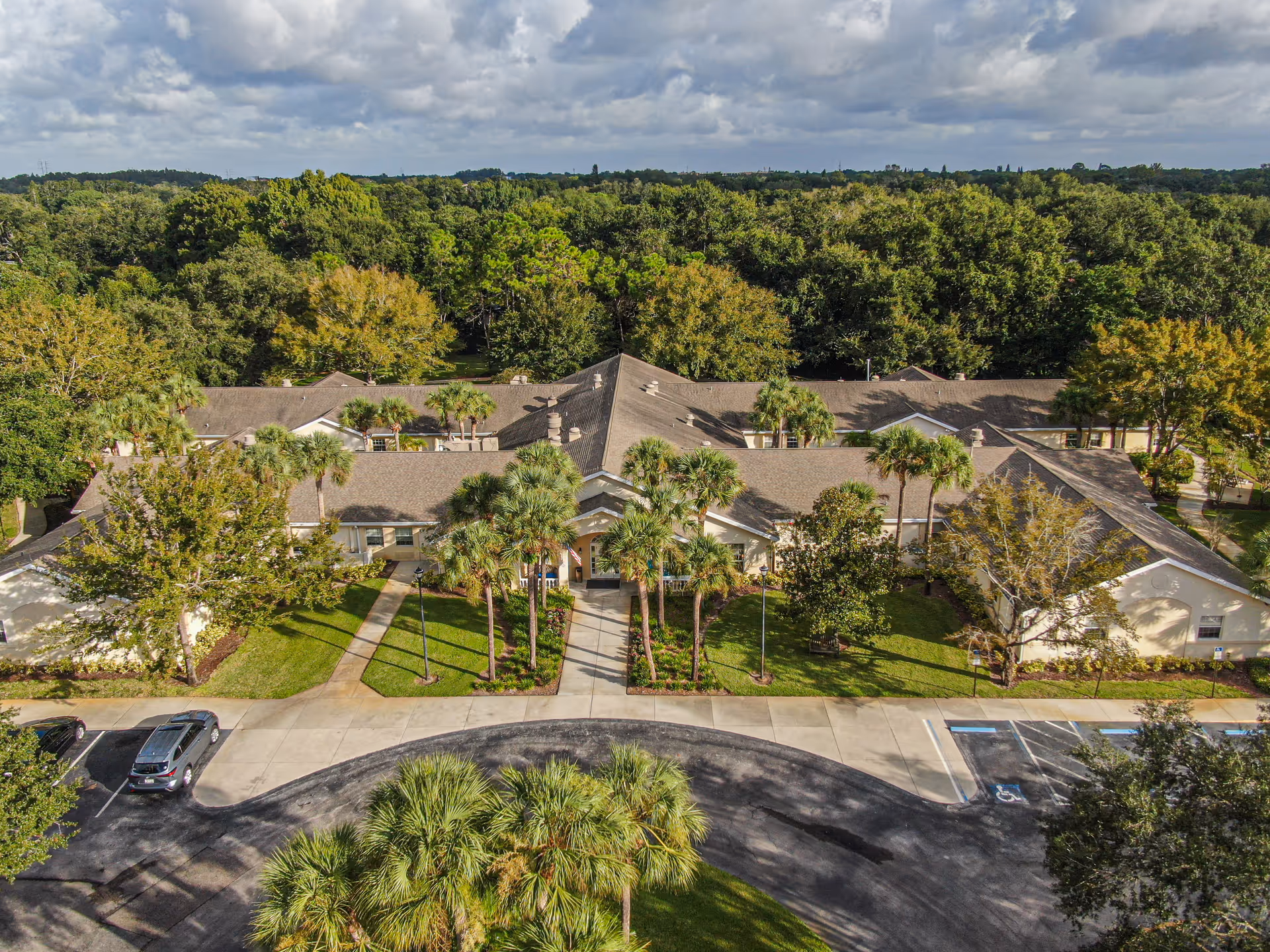 Aerial view of a single-story senior living/memory care building with palm trees, entrance drive and parking, set against a backdrop of dense trees.