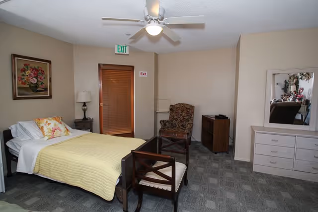 A senior care bedroom with a single bed covered with a yellow quilt and floral pillows. There is a wooden chair with a cushioned seat at the foot of the bed, a floral upholstered armchair in the corner, a wooden dresser with a mirror, and a small wooden cabinet. The room has beige walls, a ceiling fan with a light, a framed floral painting on the wall, and an exit door with an exit sign above it.