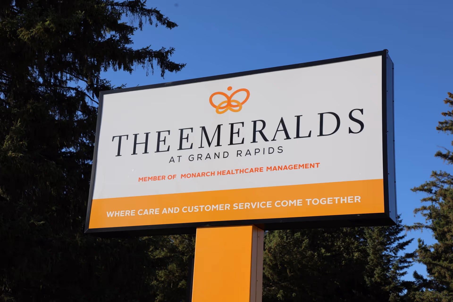 A large outdoor sign for The Emeralds at Grand Rapids, a member of Monarch Healthcare Management, with the tagline 'Where care and customer service come together'. The sign is mounted on an orange post with trees and a clear blue sky in the background.