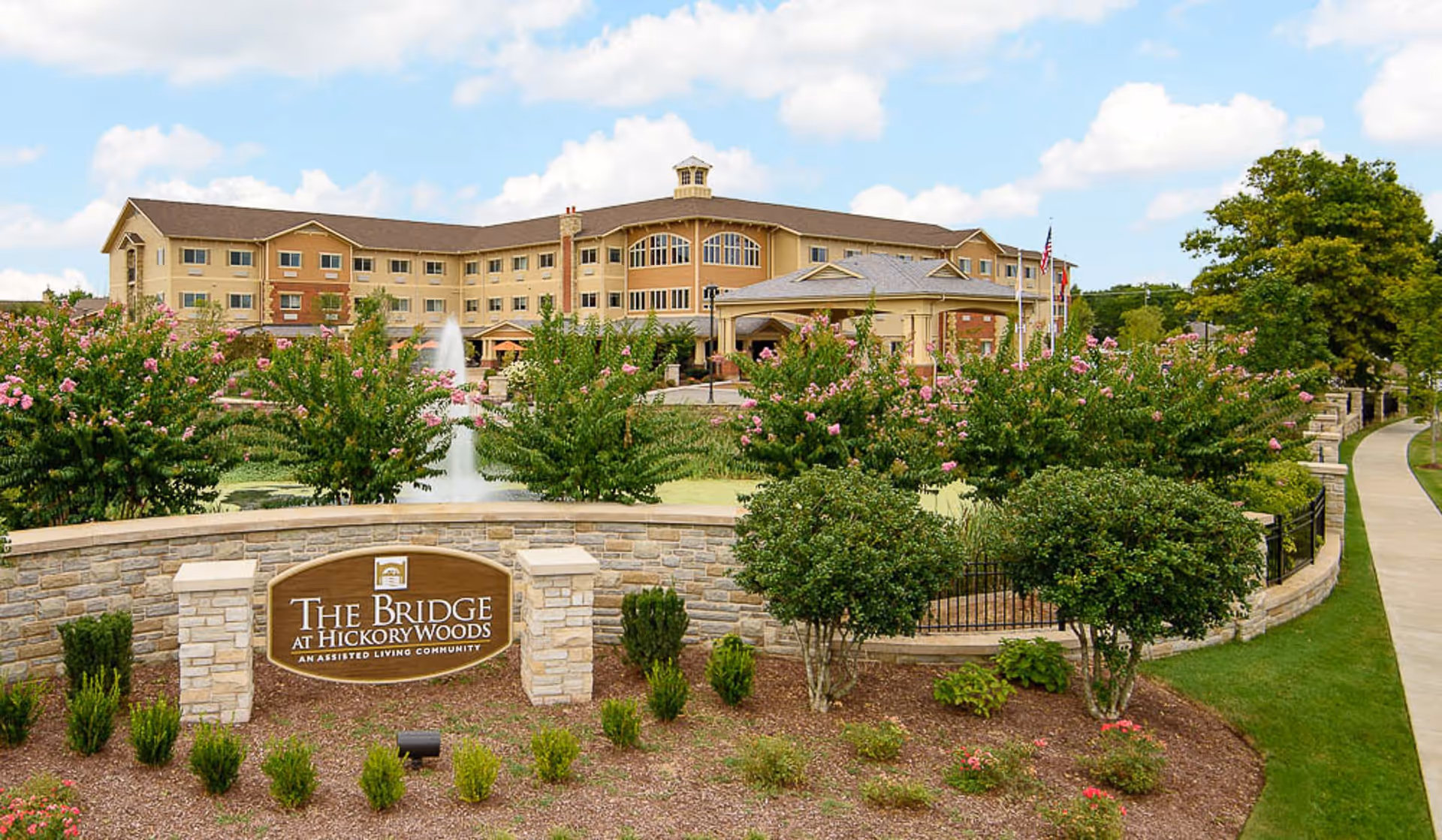 Exterior view of The Bridge at Hickory Woods assisted living community building with a landscaped garden, flowering bushes, a water fountain, and a stone sign displaying the facility's name. The sky is partly cloudy and a sidewalk runs along the right side of the image.