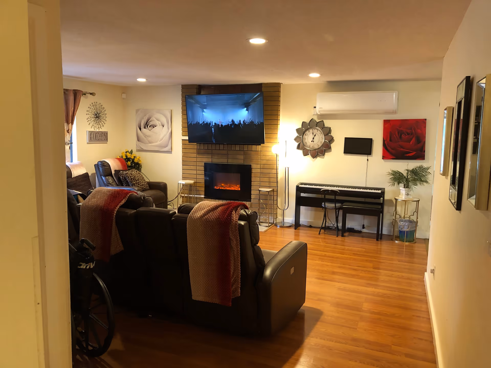 Cozy living room with reclining chairs facing a wall-mounted TV and electric fireplace, a piano against the far wall, and hardwood floors.