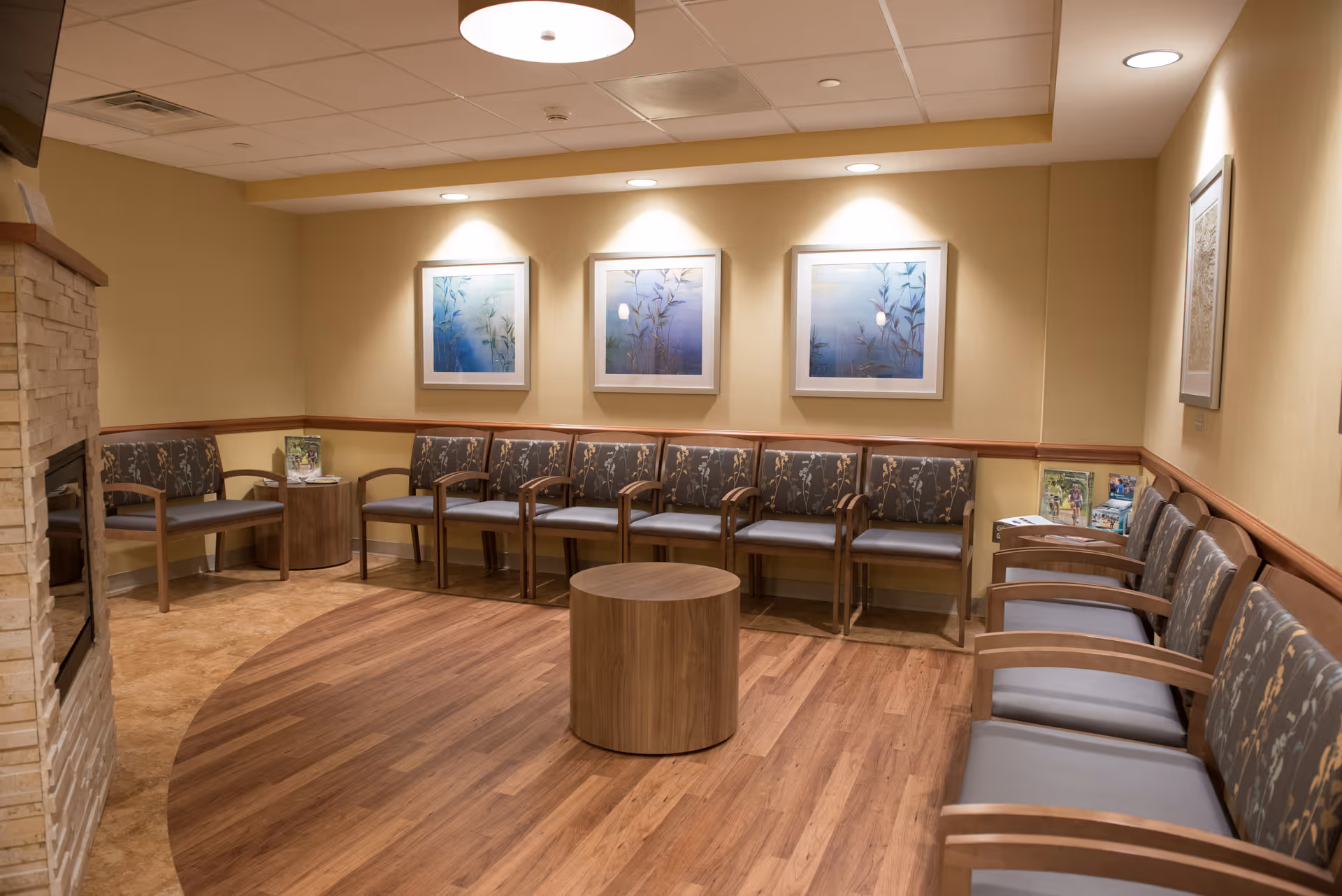 Beige-walled waiting lounge with numerous wooden-framed chairs arranged around a circular wooden table.