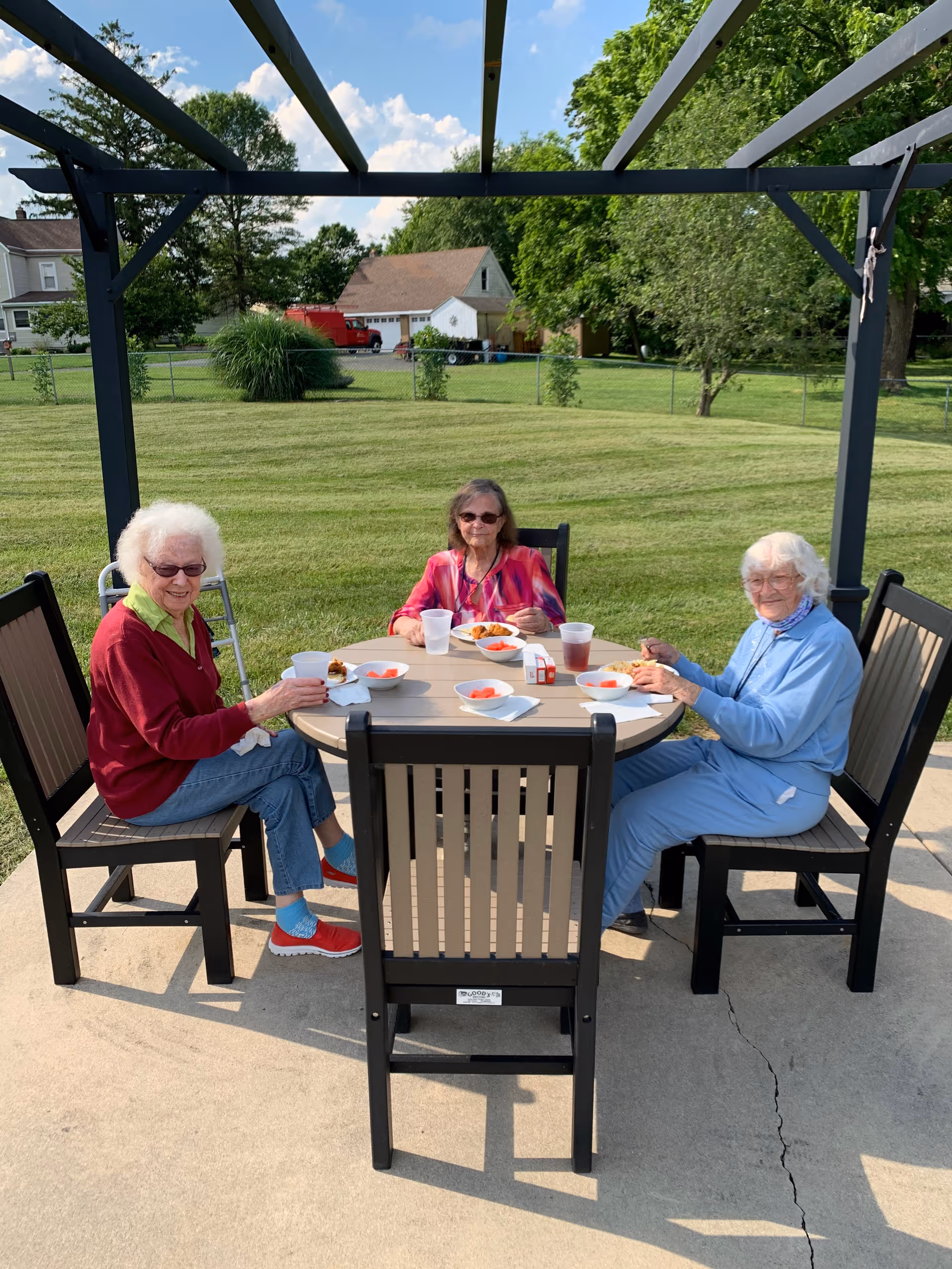 Three elderly women sit at an outdoor dining table under a pergola.