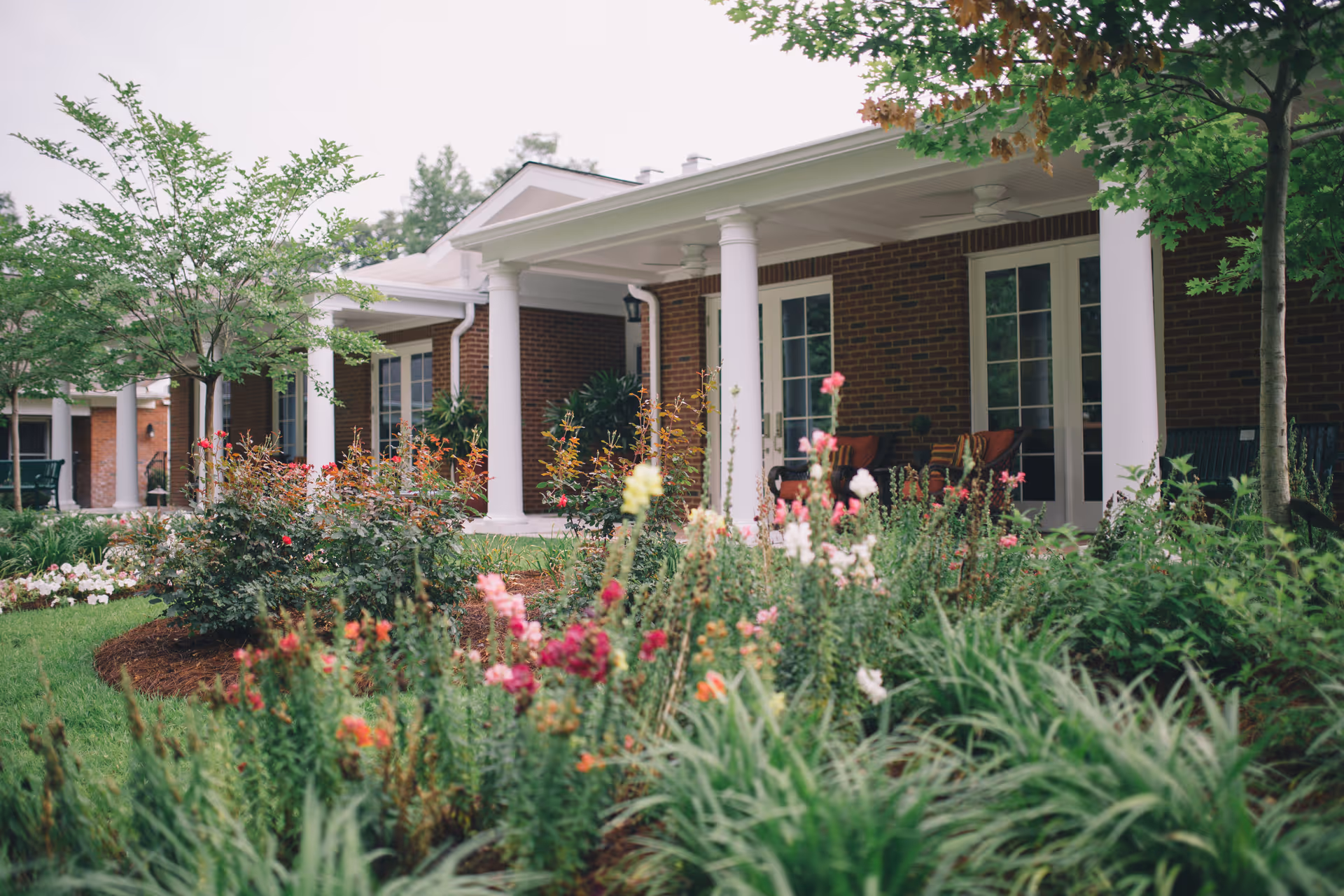 Front view of a brick hospice building with white columns and a colorful garden.