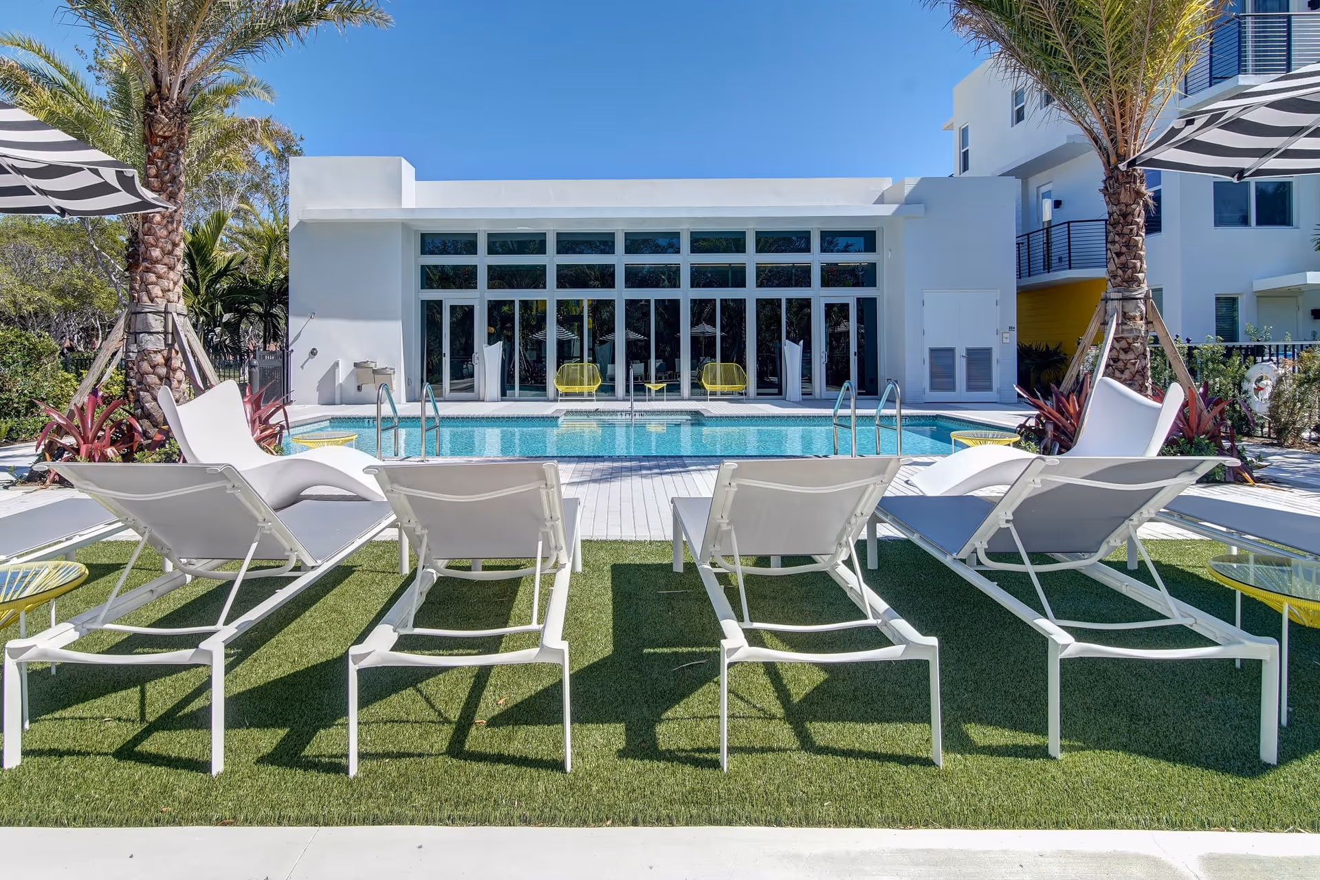 Outdoor pool area at Upside Doral facility with white lounge chairs on artificial grass, palm trees, striped umbrellas, and a modern white building with large glass windows in the background under a clear blue sky.
