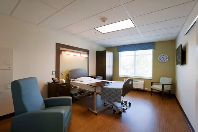 A clean and well-lit patient room in a healthcare facility featuring a hospital bed with a side table, a blue armchair, a wooden cabinet, a window with a blue valance, a wall-mounted clock, and a flat-screen TV mounted on the wall.