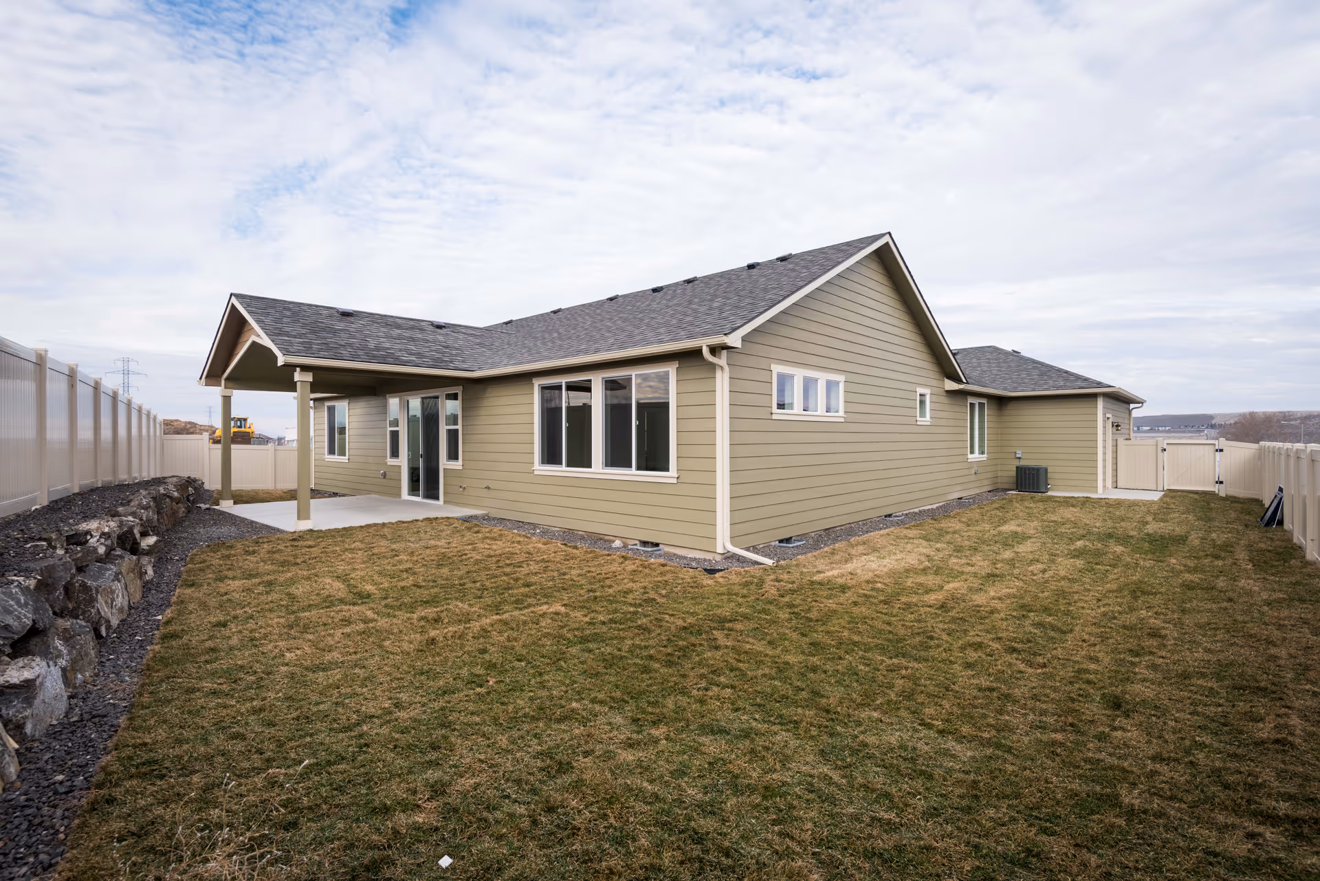Exterior view of a single-story building with beige siding and a gray shingled roof, surrounded by a fenced yard with grass and a rock border on one side under a cloudy sky.