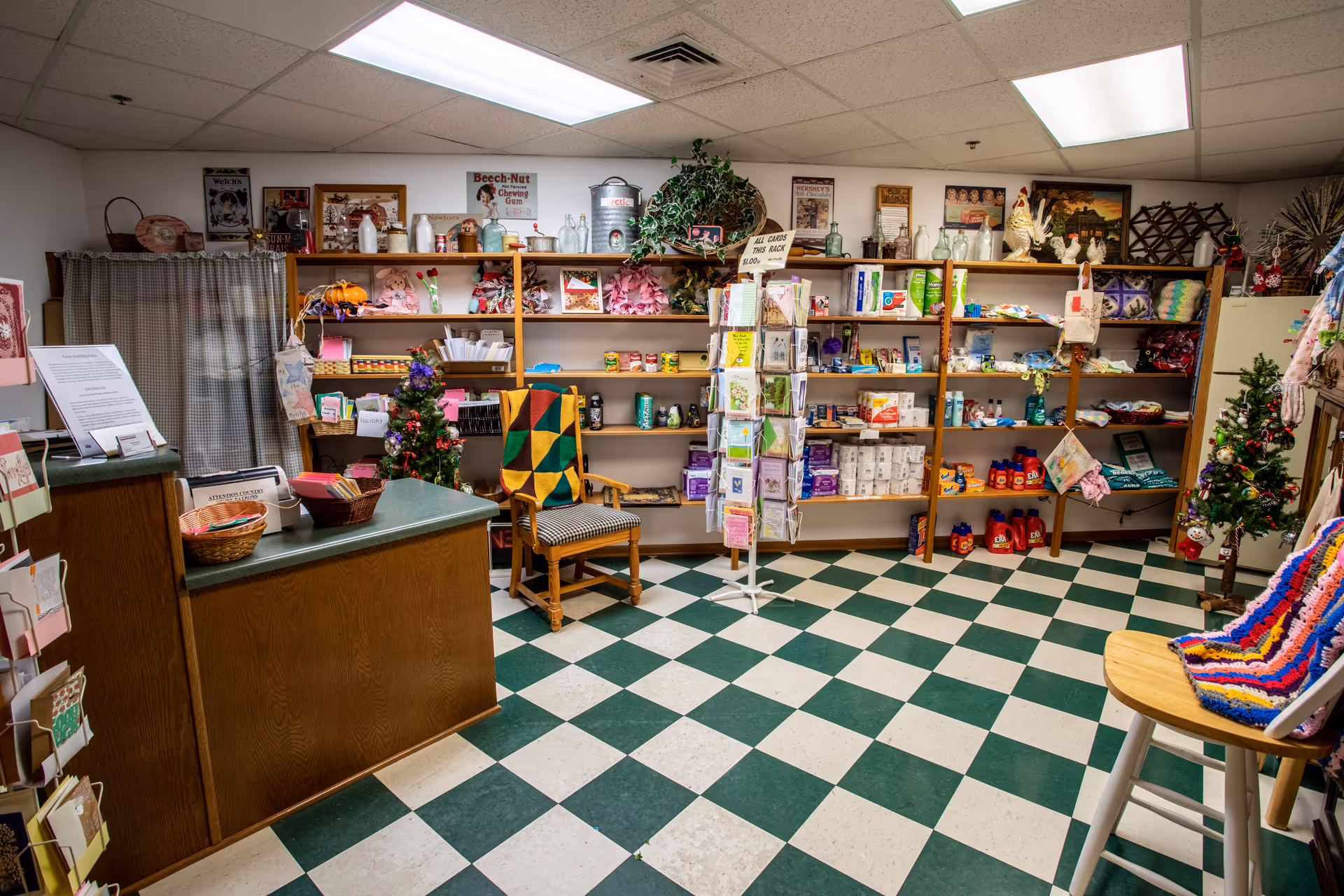 Small gift shop inside a senior living facility with shelves of toiletries and cards, a checkout counter, and a green-and-white checkered floor.