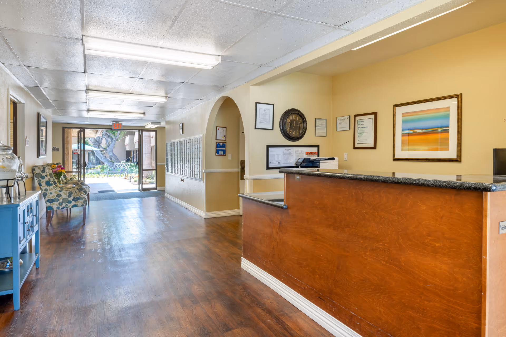 Reception area of a senior living facility with a wooden front desk on the right, a wall clock, framed certificates and artwork on the wall behind the desk. On the left side, there are two floral patterned chairs next to a blue console table with a water dispenser and flowers. The hallway leads to glass doors opening to an outdoor garden area with trees and seating.