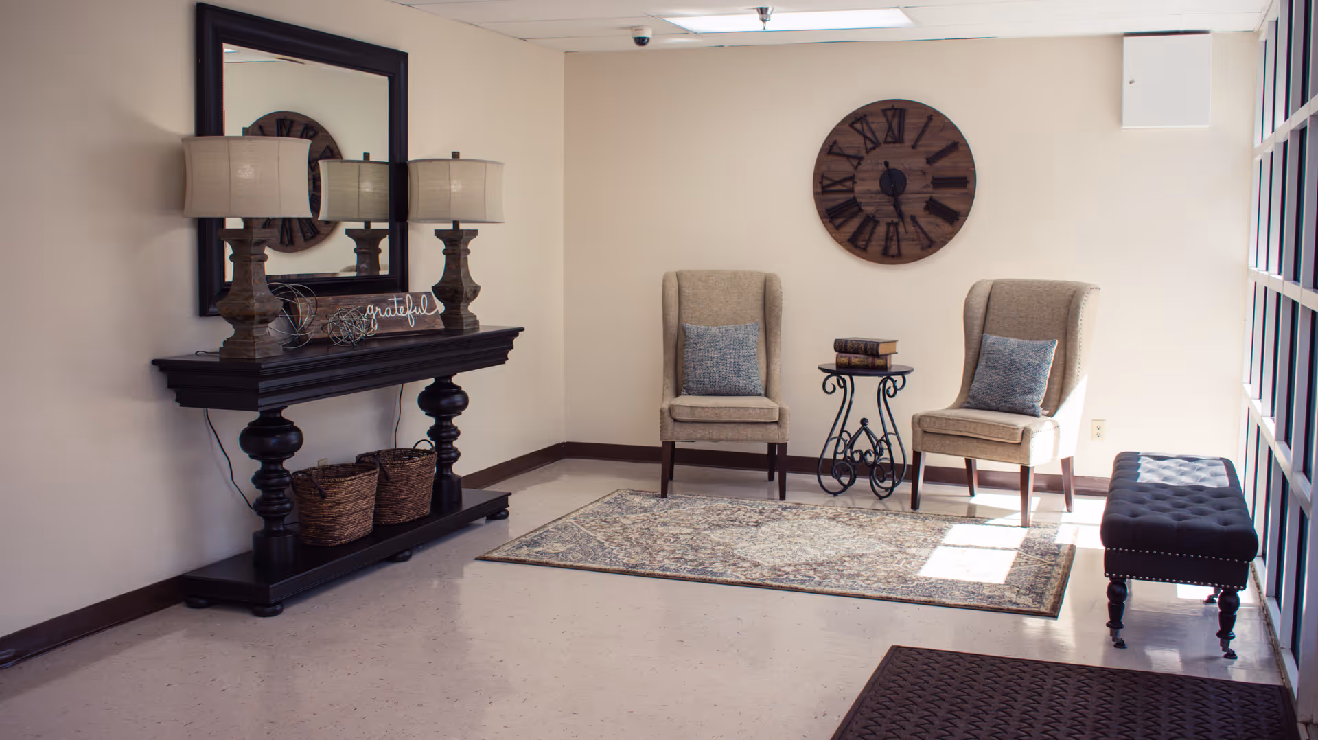 A cozy seating area with two beige armchairs with blue cushions, a small decorative table with books, a large wall clock, a dark wooden console table with two lamps and decorative items, a large mirror above the console, two wicker baskets underneath, and a cushioned bench near a window letting in natural light.