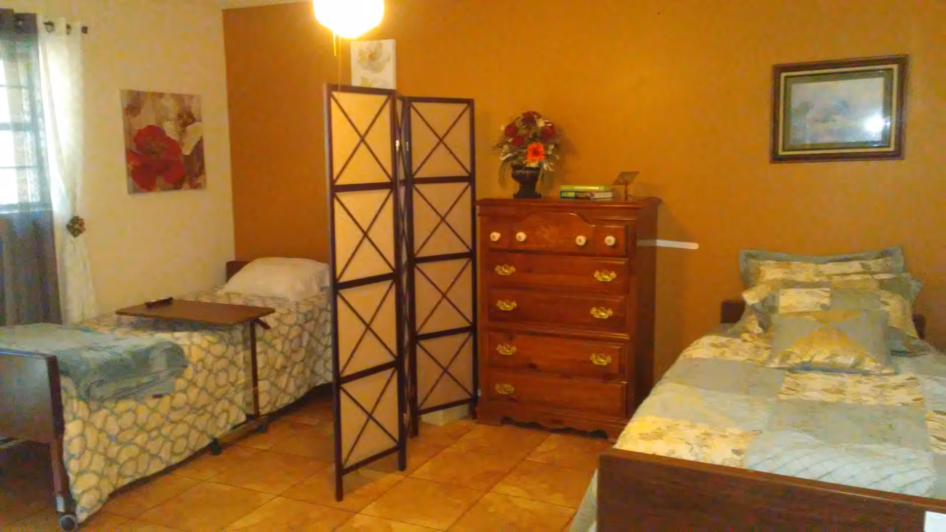 A bedroom with two single beds separated by a folding room divider. One bed has a patterned bedspread and a small overbed table, while the other bed has a patchwork quilt and multiple pillows. There is a wooden chest of drawers with a flower arrangement and books on top against a brown wall. The room has tiled flooring, a window with curtains, and framed artwork on the walls.