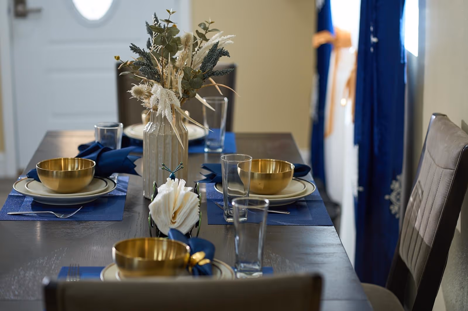 A dining table set with plates, bowls, glasses, and a dried flower centerpiece.