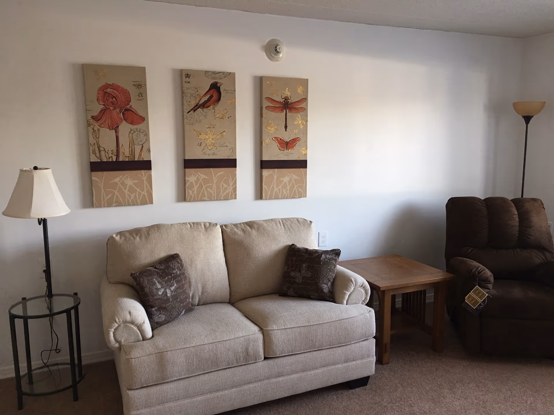A beige sofa with pillows, wooden side table, and a brown recliner in a living room.