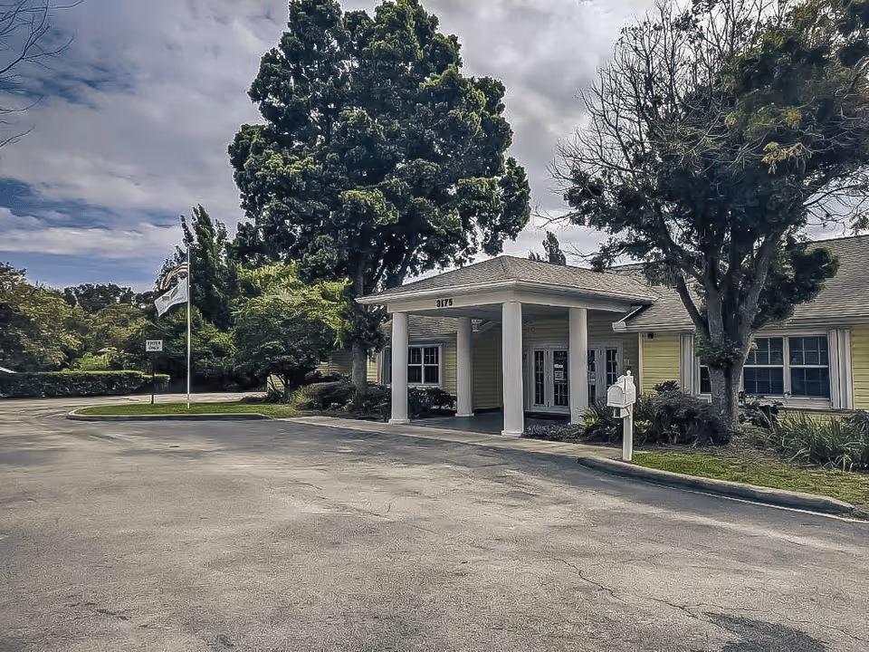 Exterior view of a single-story senior living facility building with a covered entrance supported by white columns, surrounded by trees and greenery under a partly cloudy sky.