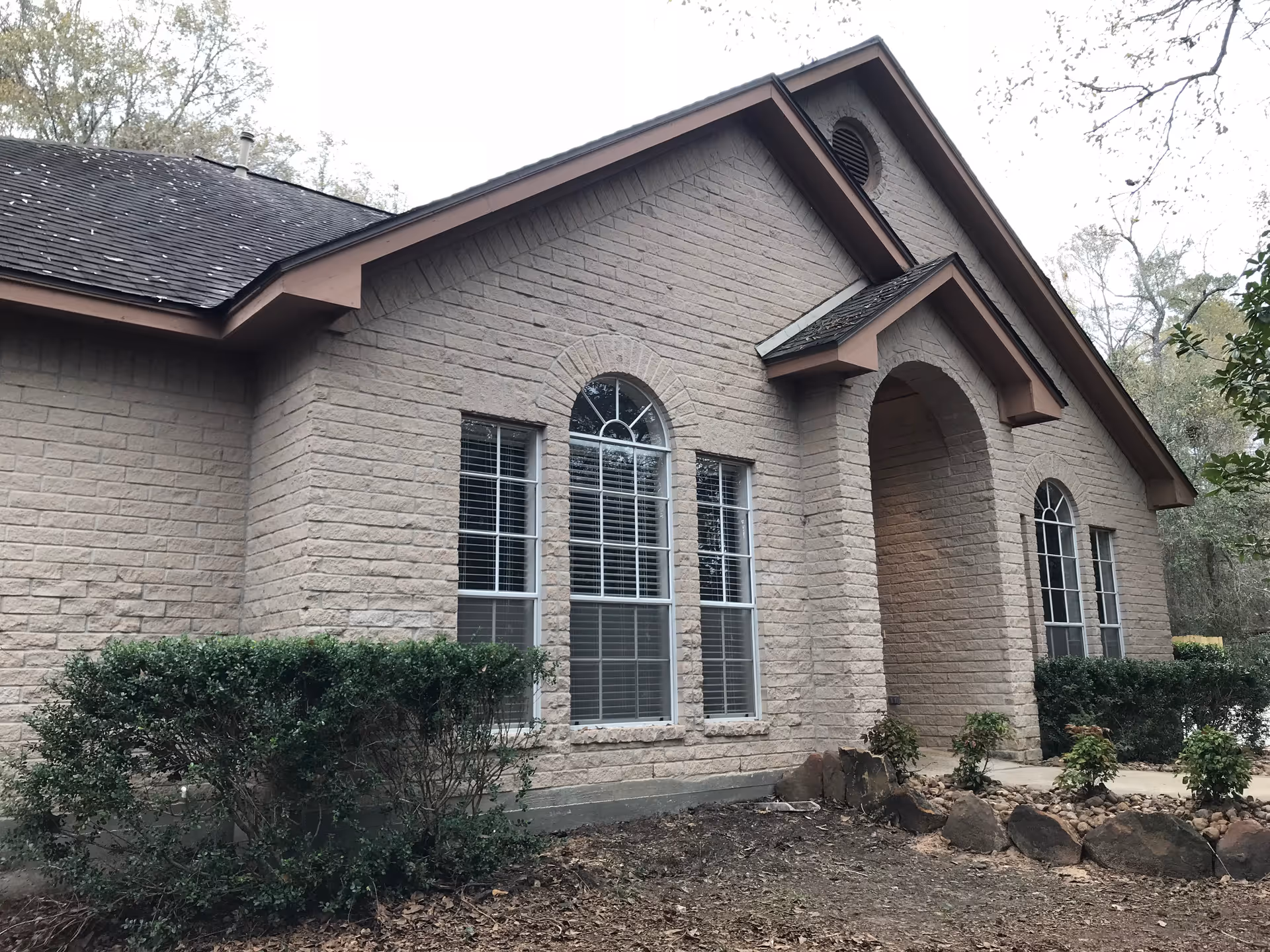 Exterior view of a single-story brick building with arched windows and an arched entrance. There are shrubs and small plants along the front of the building, and trees are visible in the background.