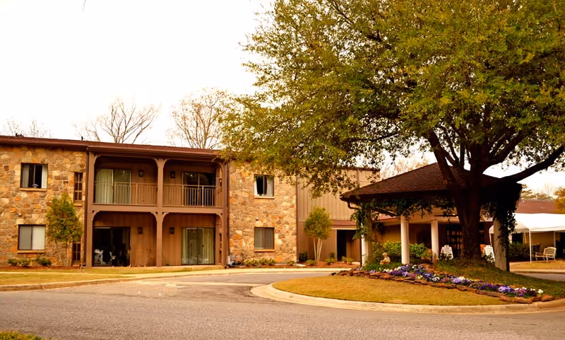Two-story stone-faced senior living building with a covered entrance, circular driveway, large tree, and flowerbed in front.