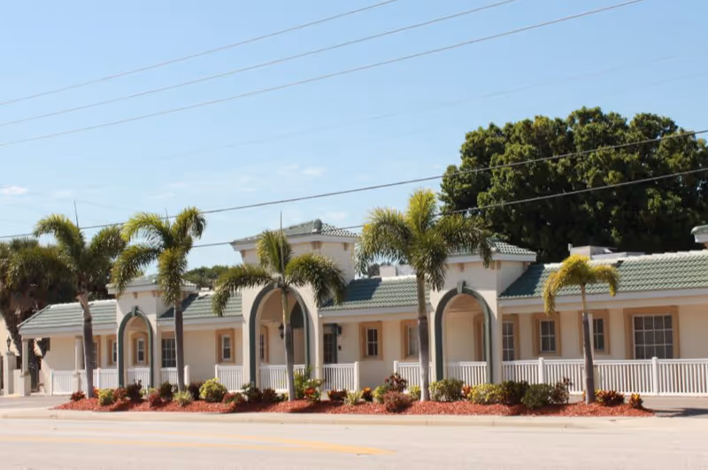 Exterior view of a single-story building with a green tiled roof, white walls, and multiple arched entryways. The front is lined with palm trees and landscaped with red mulch and various plants. The sky is clear and blue.