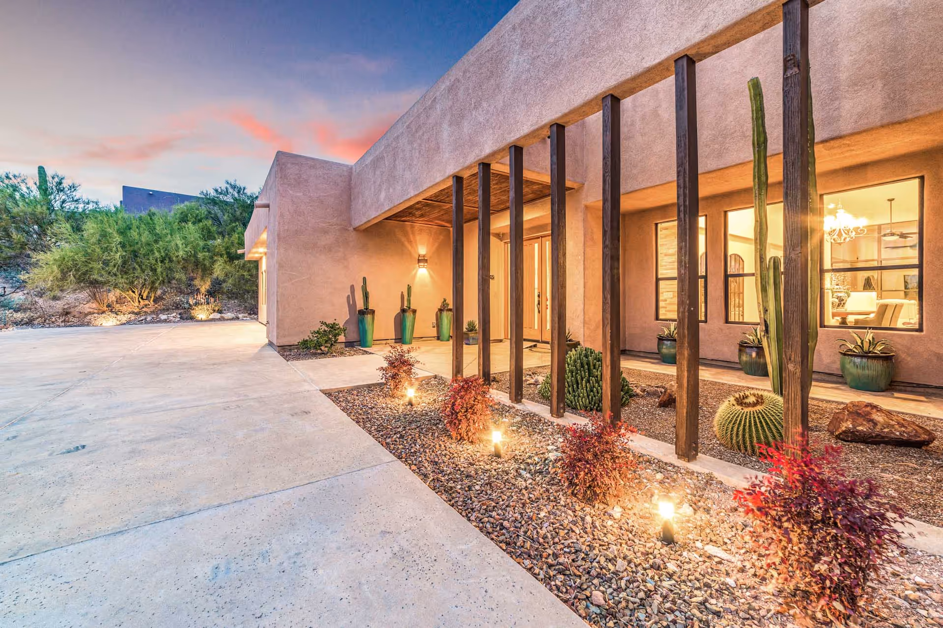 Exterior view of Desert Gardens Assisted Living and Memory Care facility at dusk, showing a modern building with desert landscaping including cacti and small shrubs, illuminated pathway lights, and large windows revealing a warmly lit interior.