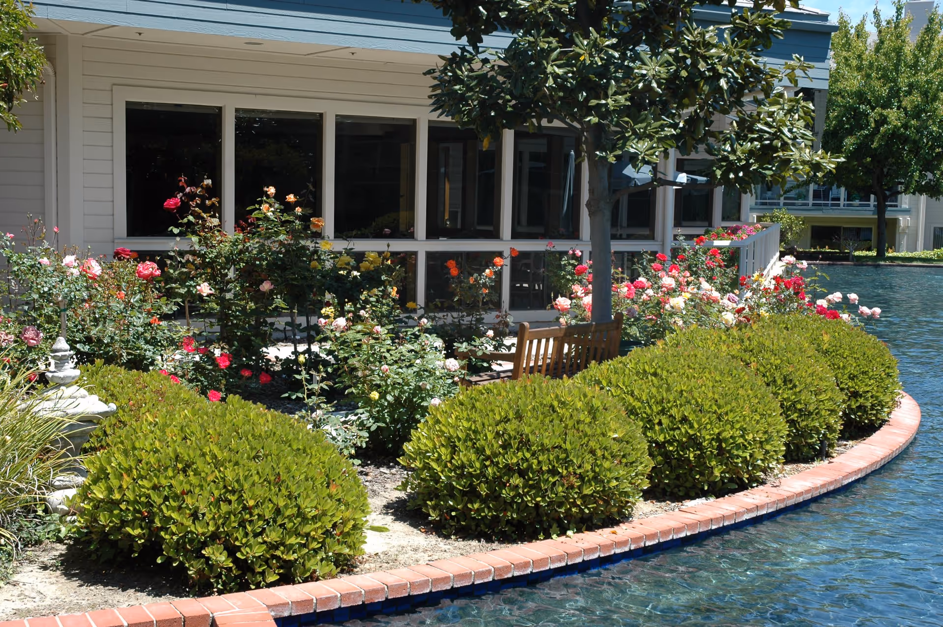 A landscaped garden area with neatly trimmed bushes and colorful blooming flowers along the edge of a curved brick border next to a clear water body. A wooden bench is partially visible behind the bushes, and a building with large windows is in the background.