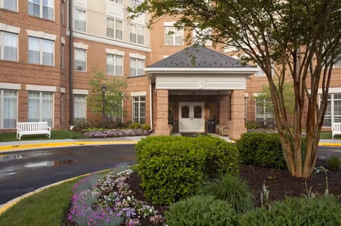 Entrance area of a senior living facility with a covered drop-off zone, surrounded by well-maintained landscaping including bushes, flowers, and trees. The building is made of brick with multiple windows and white benches are visible on either side of the entrance.