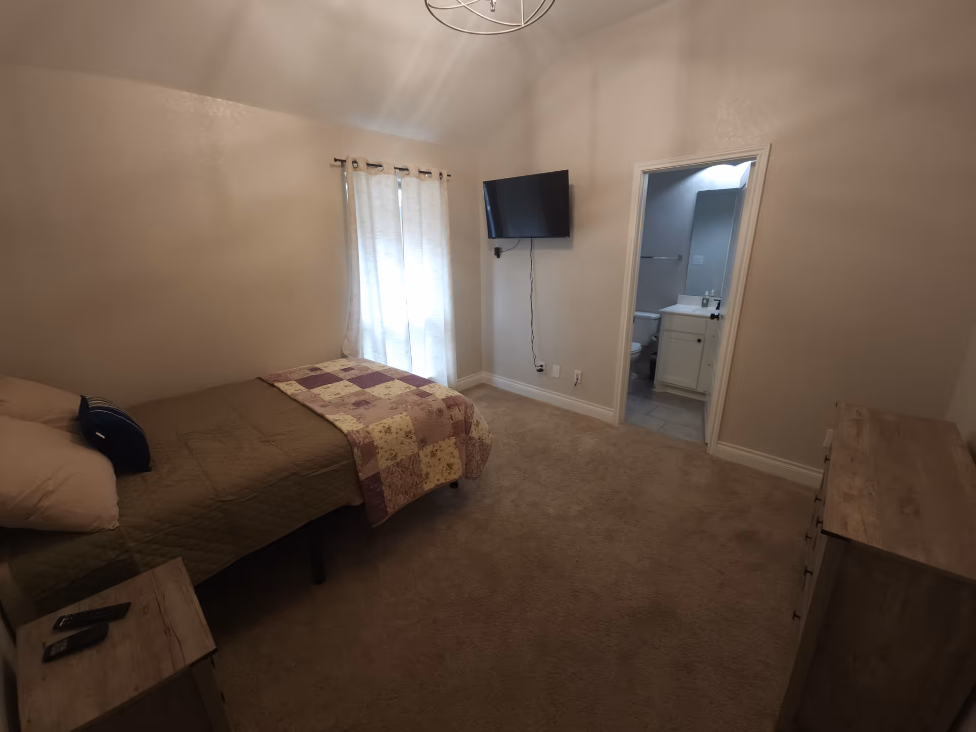 Neutral bedroom with a bed, dresser, wall-mounted TV, and bathroom door.