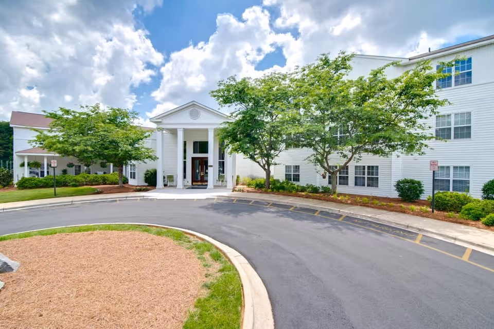 White three-story building with a columned main entrance, trees, landscaping, and a curved driveway.