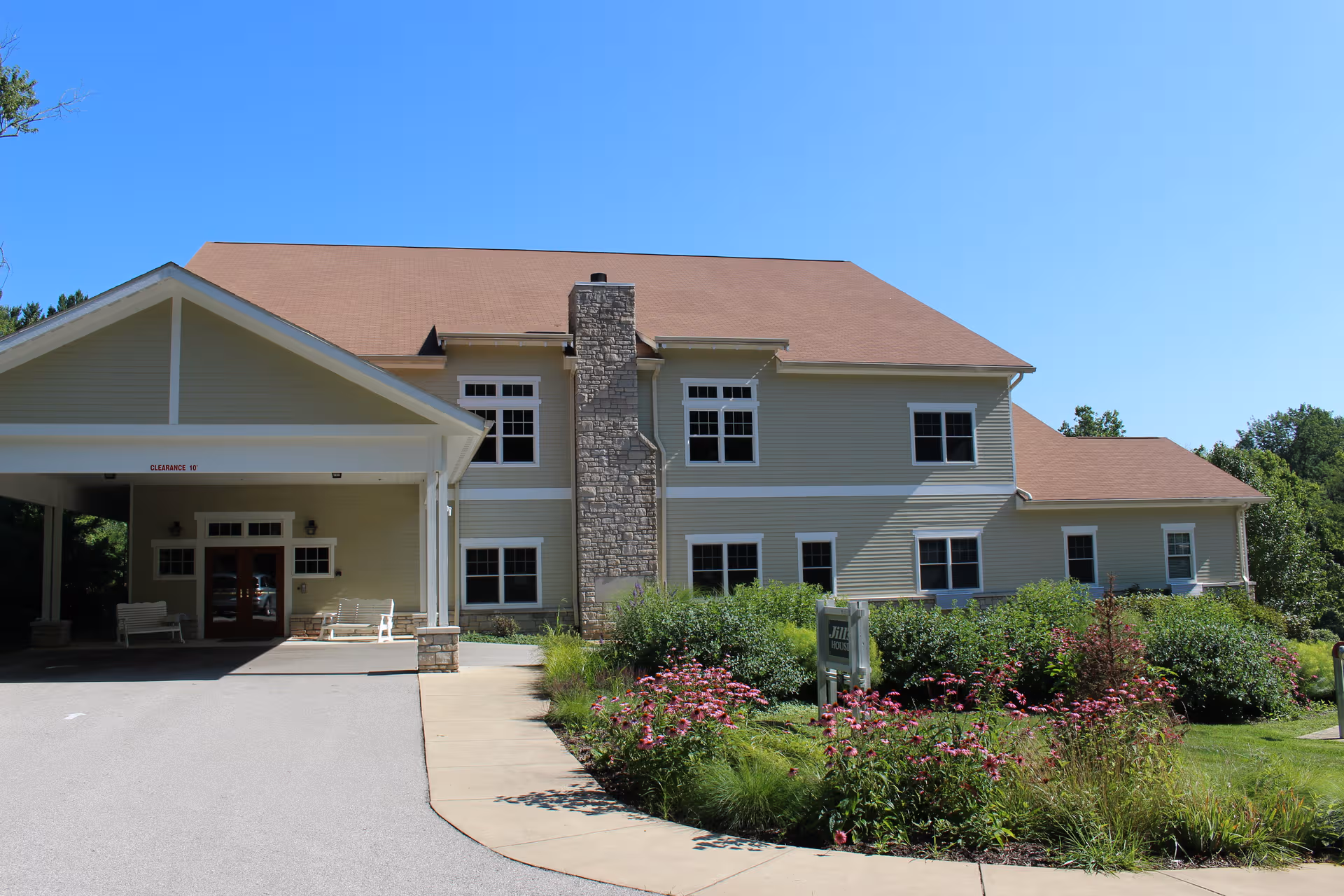 Two-story beige memory care facility with stone chimney and a covered entrance, surrounded by flowers.