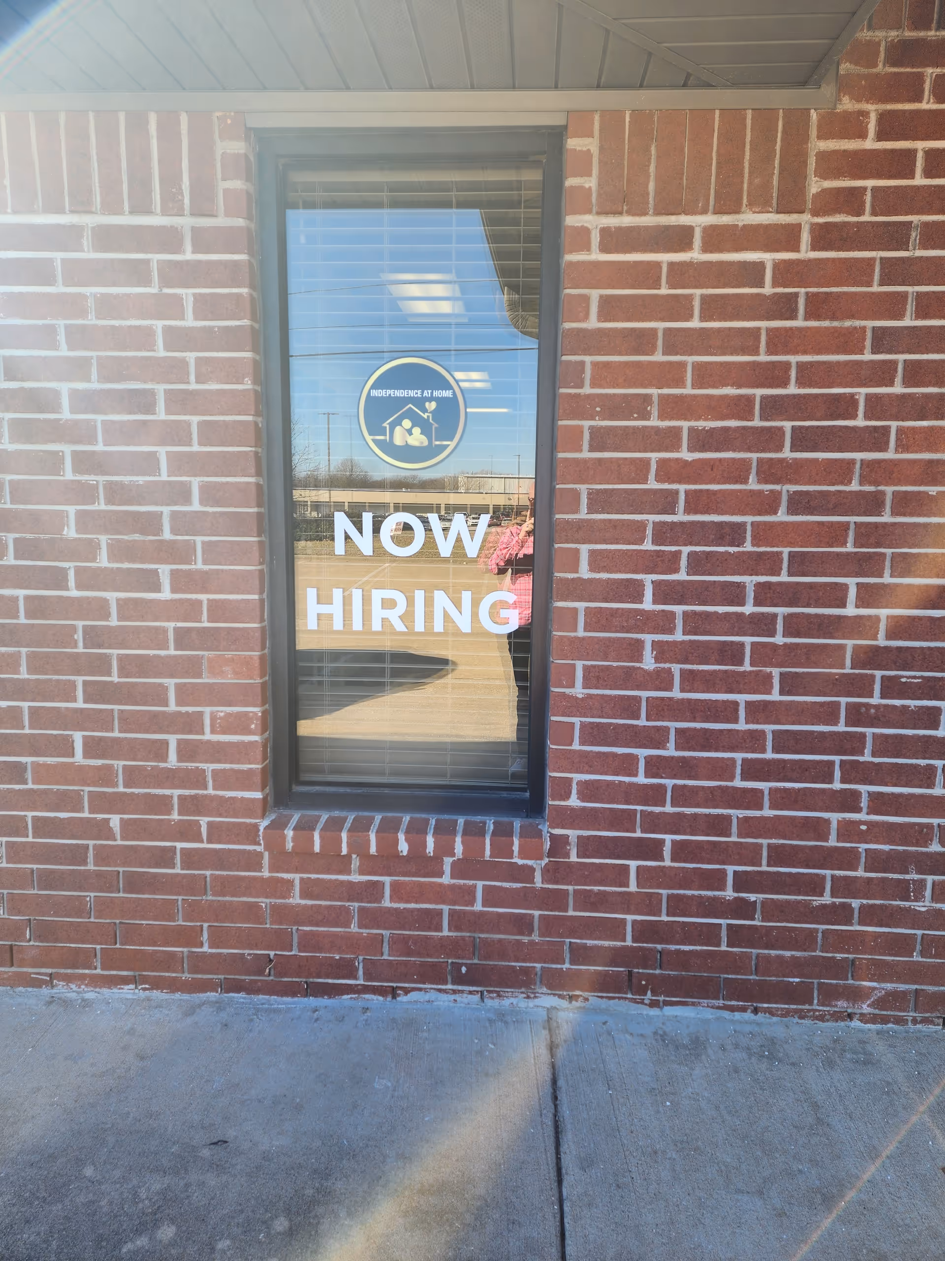 Brick building exterior with a window displaying Independence at Home logo and NOW HIRING sign.