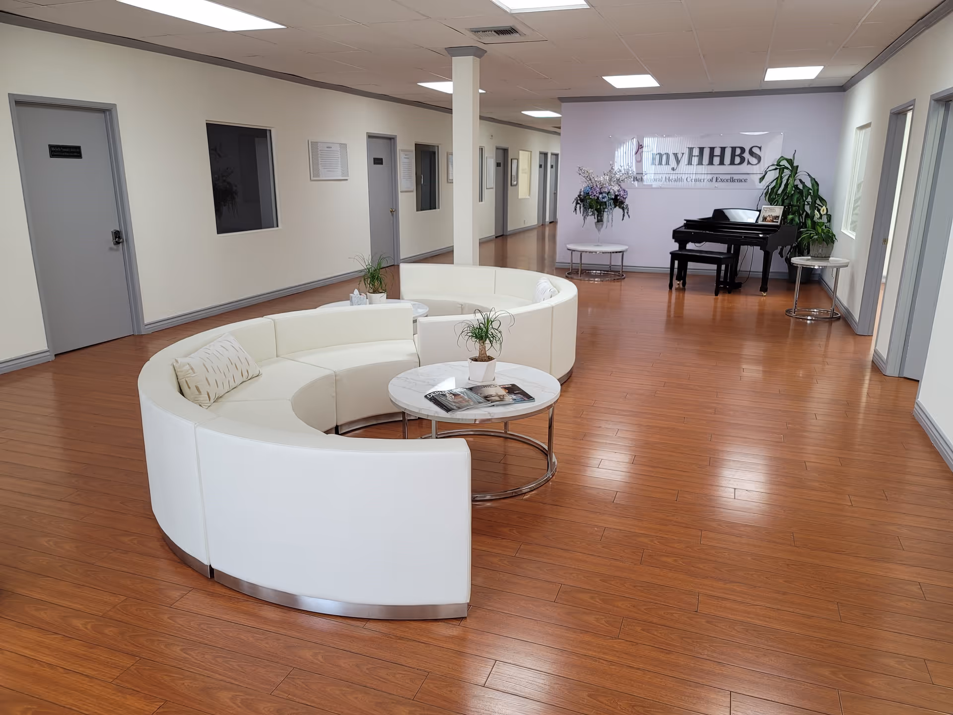 Modern facility lobby featuring curved white seating, round coffee tables, a grand piano, and plants.