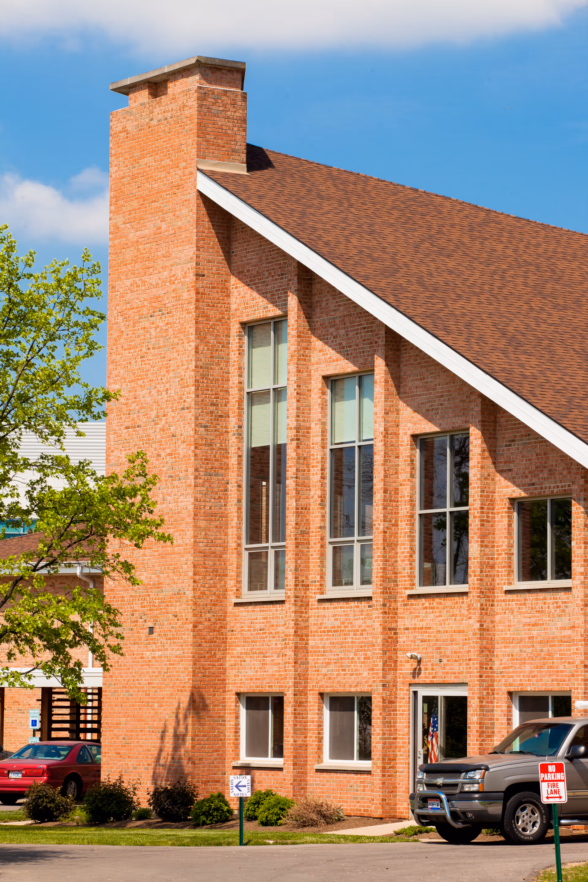Exterior view of a brick building with large vertical windows and a steeply sloped roof under a blue sky. There are a few parked cars and a tree with green leaves near the building.
