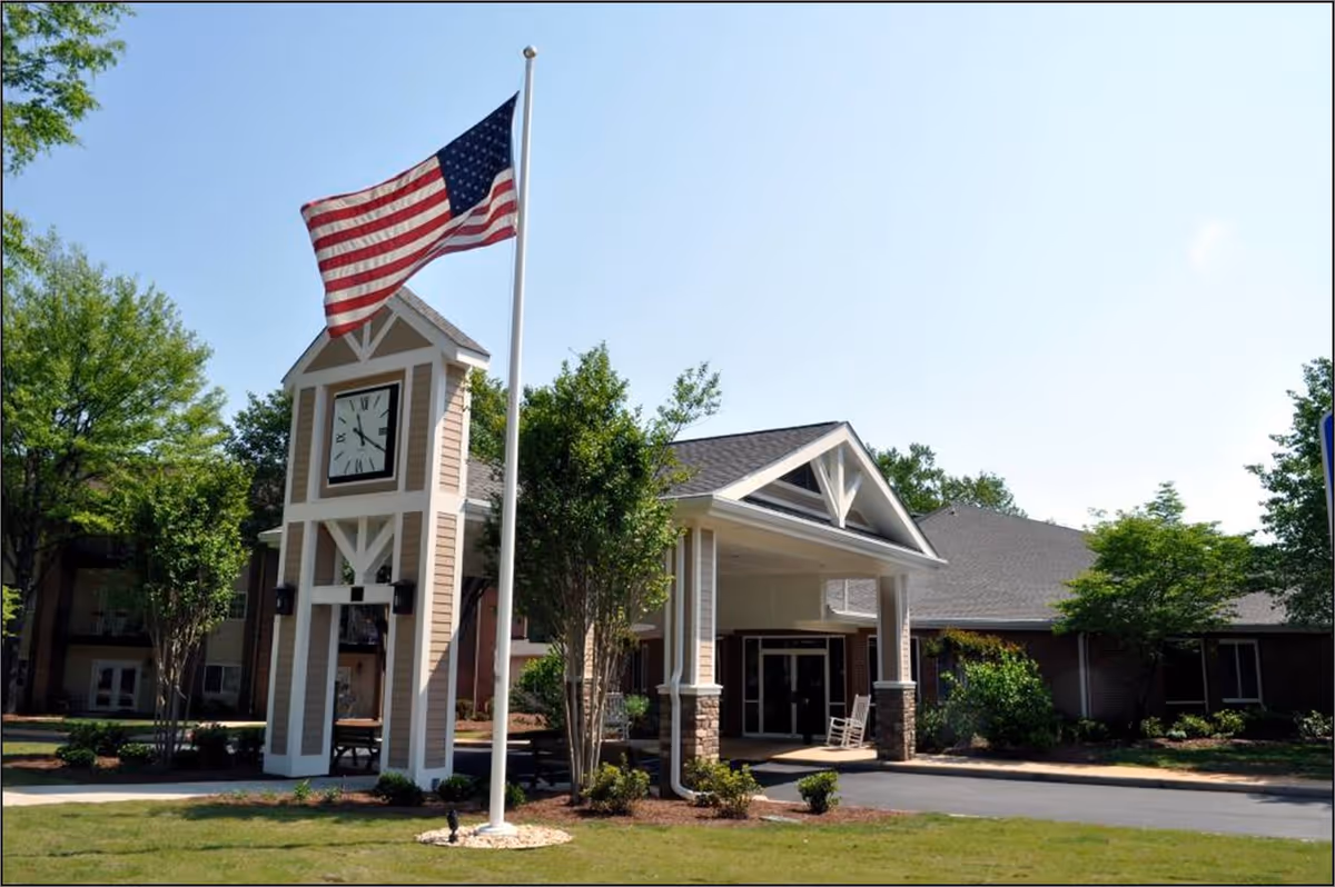 Exterior view of a senior living facility with a clock tower and an American flag on a flagpole in front. The building has a covered entrance with a rocking chair and is surrounded by trees and landscaping under a clear blue sky.