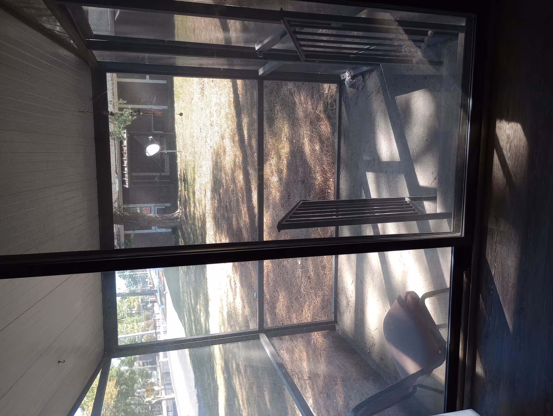 Sunroom with glass walls, metal frame, and a chair; outside lawn and trees visible.