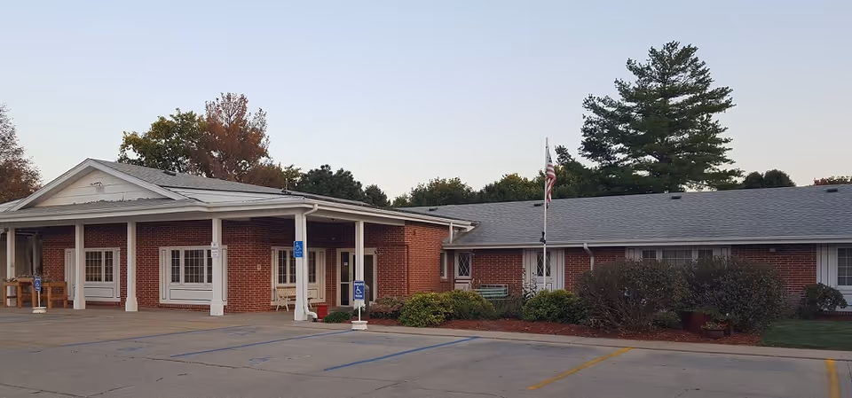 Exterior view of a single-story brick building with a covered entrance, several windows, and an American flag on a flagpole in front. There are parking spaces, including handicapped spots, in the foreground and some trees in the background.