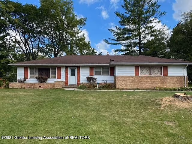 Single-story ranch-style house with brick and white siding, red shutters, a front lawn, and trees under a blue sky.