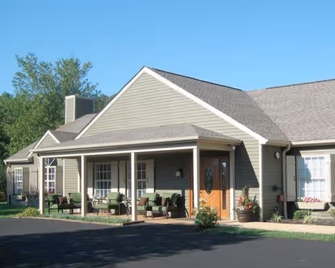 Front exterior of a single-story assisted living building with a covered porch furnished with chairs and a glass entry door.